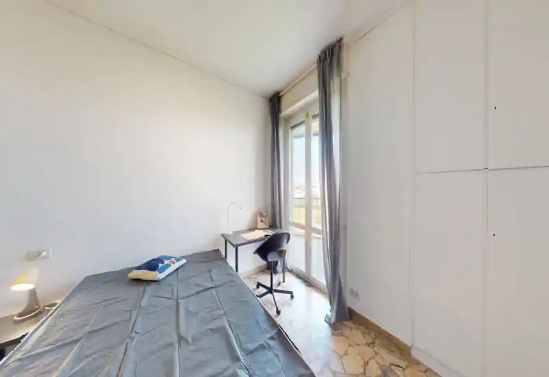 Camera singola luminosa e ordinata con letto a sinistra, una scrivania vicino alla finestra grande e armadi a muro. La luce naturale rende l'ambiente ideale per studiare e rilassarsi.