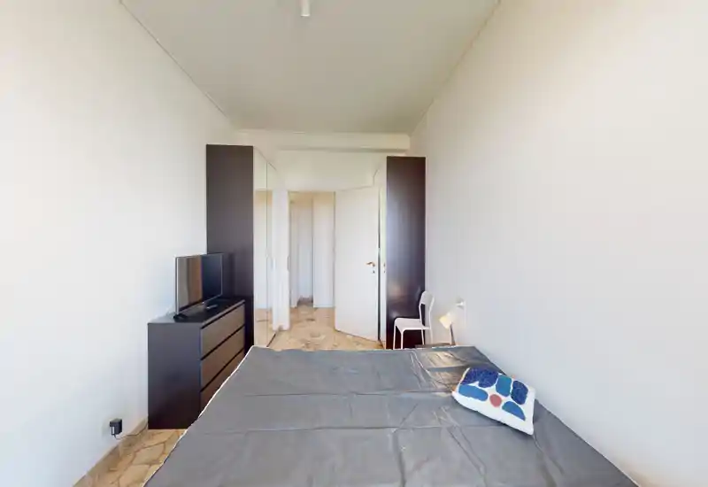 Camera da letto luminosa e minimalista con letto matrimoniale in primo piano, comò con TV e armadi alti vicino alla porta. Linee pulite e toni neutri creano un ambiente tranquillo ideale per annunci.