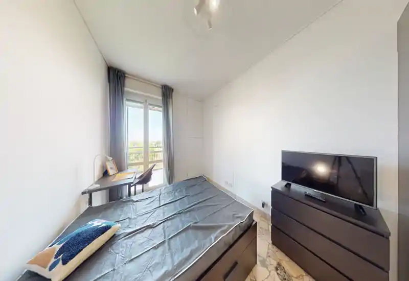 Camera da letto luminosa e moderna con letto matrimoniale, comò con TV e una piccola scrivania vicino alla finestra che lascia entrare luce naturale.