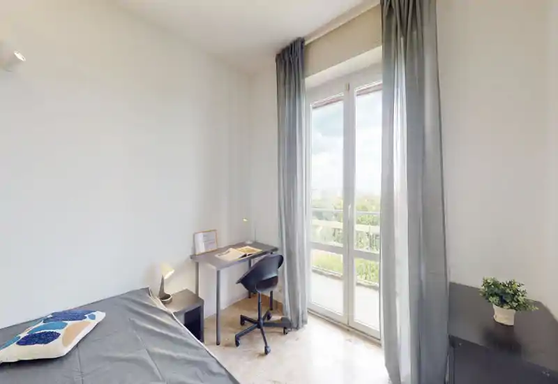 Bright, minimal single bedroom with a bed, bedside lamp, workspace and large balcony window providing abundant natural light and a pleasant city view.