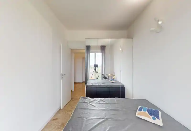 Minimal bright bedroom with a double bed, mirrored wardrobe and natural light from a window. Simple, functional styling with a bedside lamp and cushion.