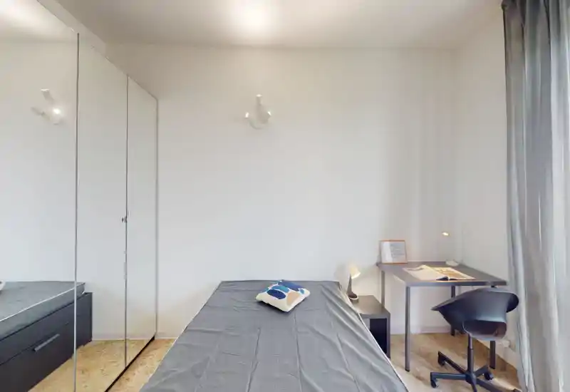 Bright, minimal bedroom with a centrally placed single bed, mirrored wardrobe and a compact study corner — clean lines and soft natural light make it comfortable for a short stay.