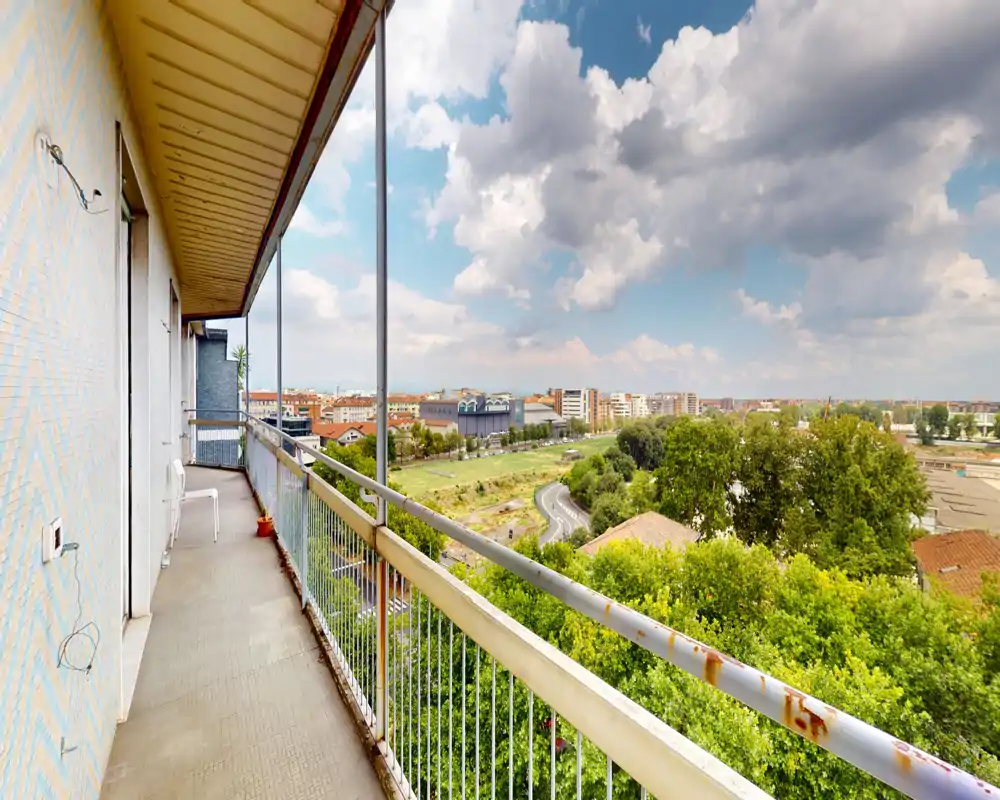 Ampio balcone con corridoio lungo e ringhiera in metallo che offre una vista panoramica su chiome e città; cielo luminoso e edifici in lontananza donano un’atmosfera ariosa.