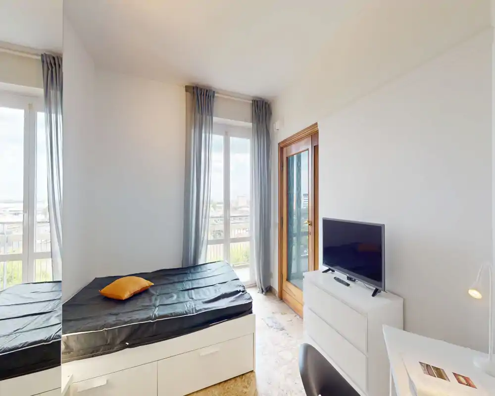 Camera da letto luminosa e compatta con letto singolo sotto grandi finestre, cassettiera con TV e scrivania — ideale per una persona che cerca molta luce naturale.