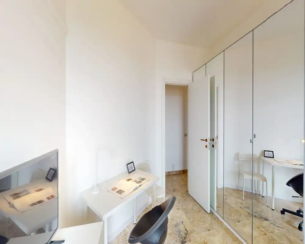 Angolo letto luminoso e minimale con scrivania, sedia e ampia armadiatura con ante specchiate. Lo spazio è pulito e arioso, adatto a una singola persona o studente.