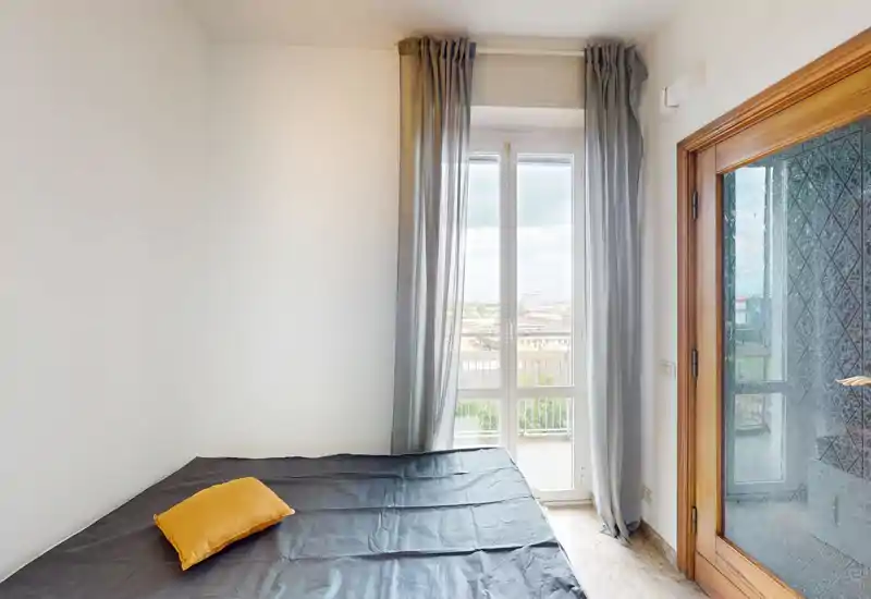 Camera da letto luminosa e minimal con letto matrimoniale coperto da una coperta scura e un cuscino giallo, ampia finestra con tende grigie che inquadra la vista sulla città.