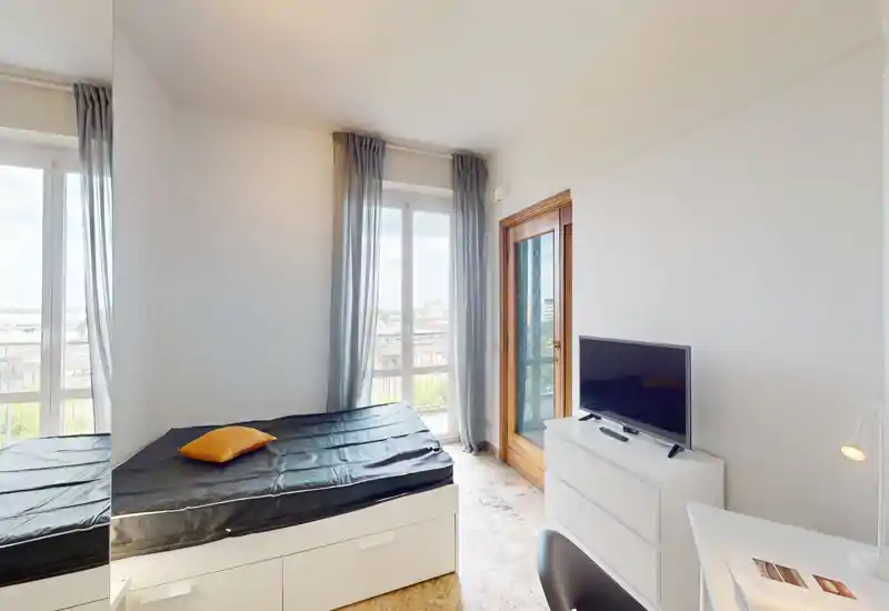 Camera da letto luminosa e compatta con letto singolo sotto grandi finestre, cassettiera con TV e scrivania — ideale per una persona che cerca molta luce naturale.