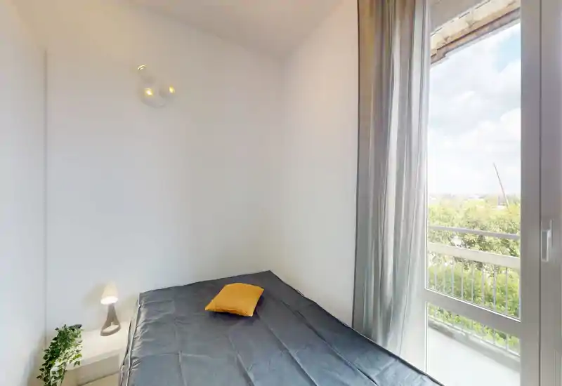 Camera da letto piccola e luminosa con letto matrimoniale in biancheria scura, cuscino giallo come dettaglio, abat-jour e porta sul balcone che illumina la stanza.