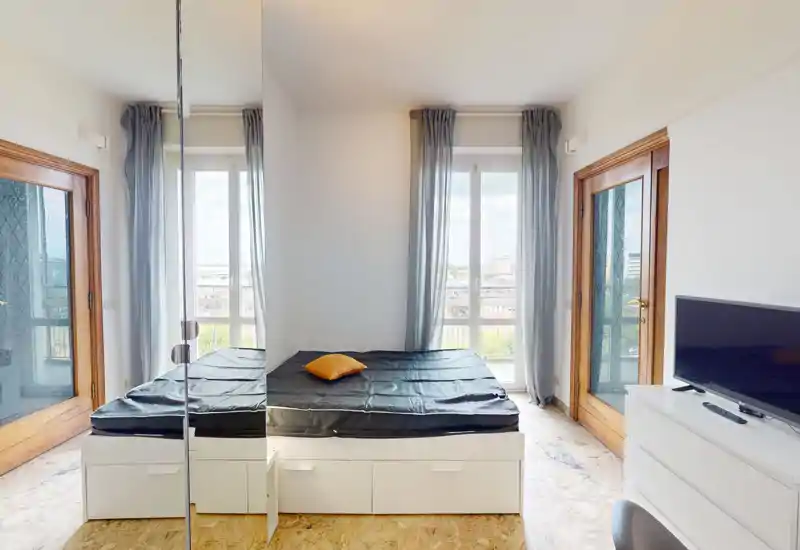 Camera da letto luminosa e minimale con letto matrimoniale centrale su struttura contenitore bianca, ampie finestre con tende leggere e un armadio con ante a specchio che amplifica lo spazio.