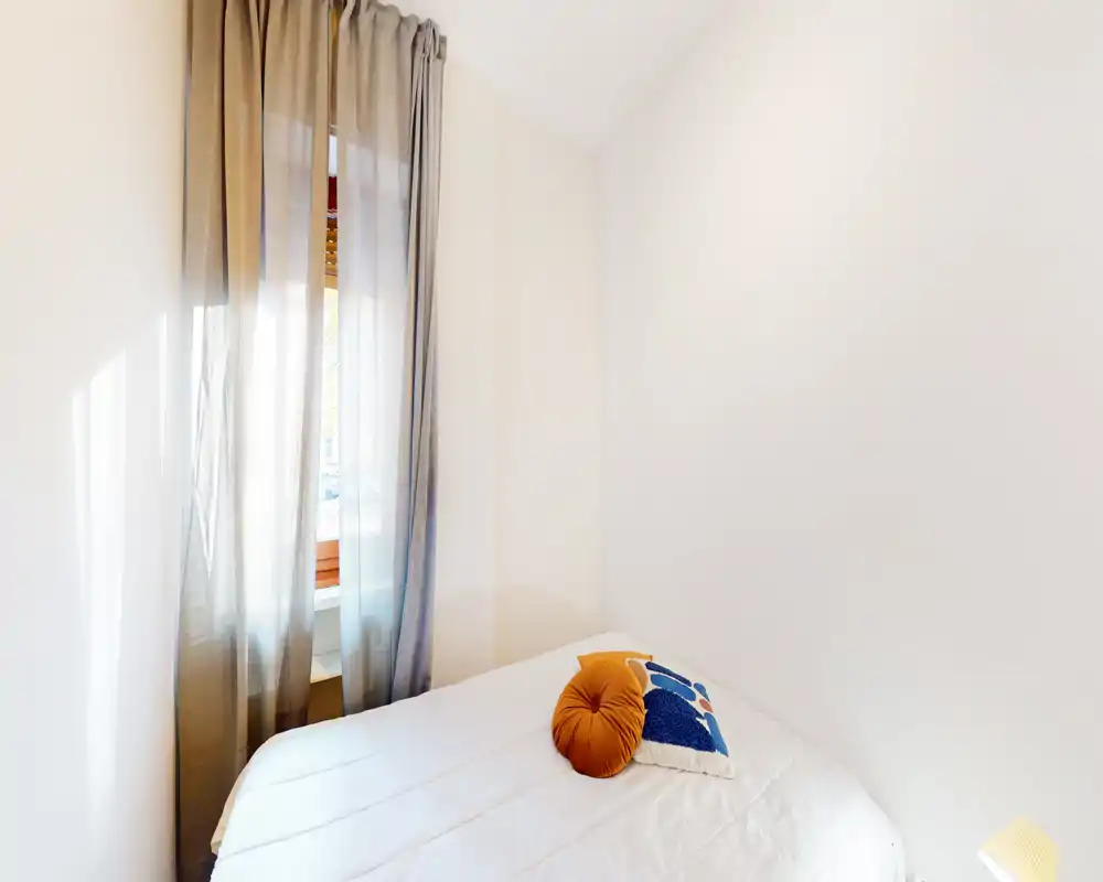 Small bright bedroom with a neatly made bed, decorative throw pillows and a sheer curtained window providing soft natural light — ideal for a cozy single or guest room.