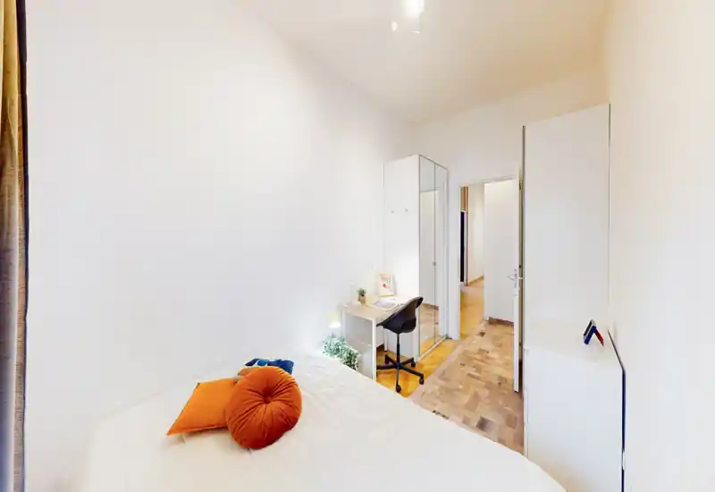 Bright, compact bedroom with a neatly made bed accented by orange cushions, a small desk and wardrobe—ideal for a single occupant or student.