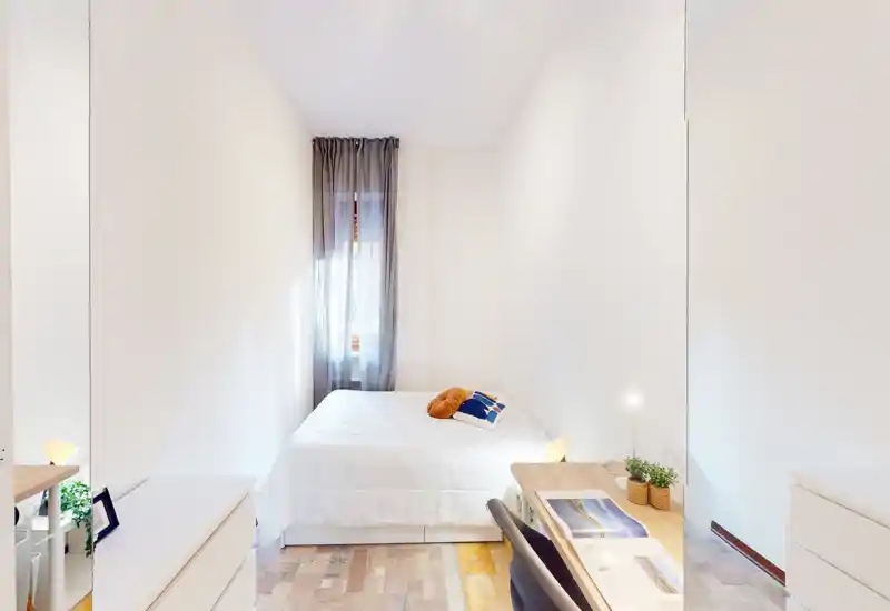 Bright, narrow bedroom with a neatly made double bed, soft natural light from a window with sheer curtains, a small desk and chair at the foot of the bed, and minimal decorative cushions and plants.