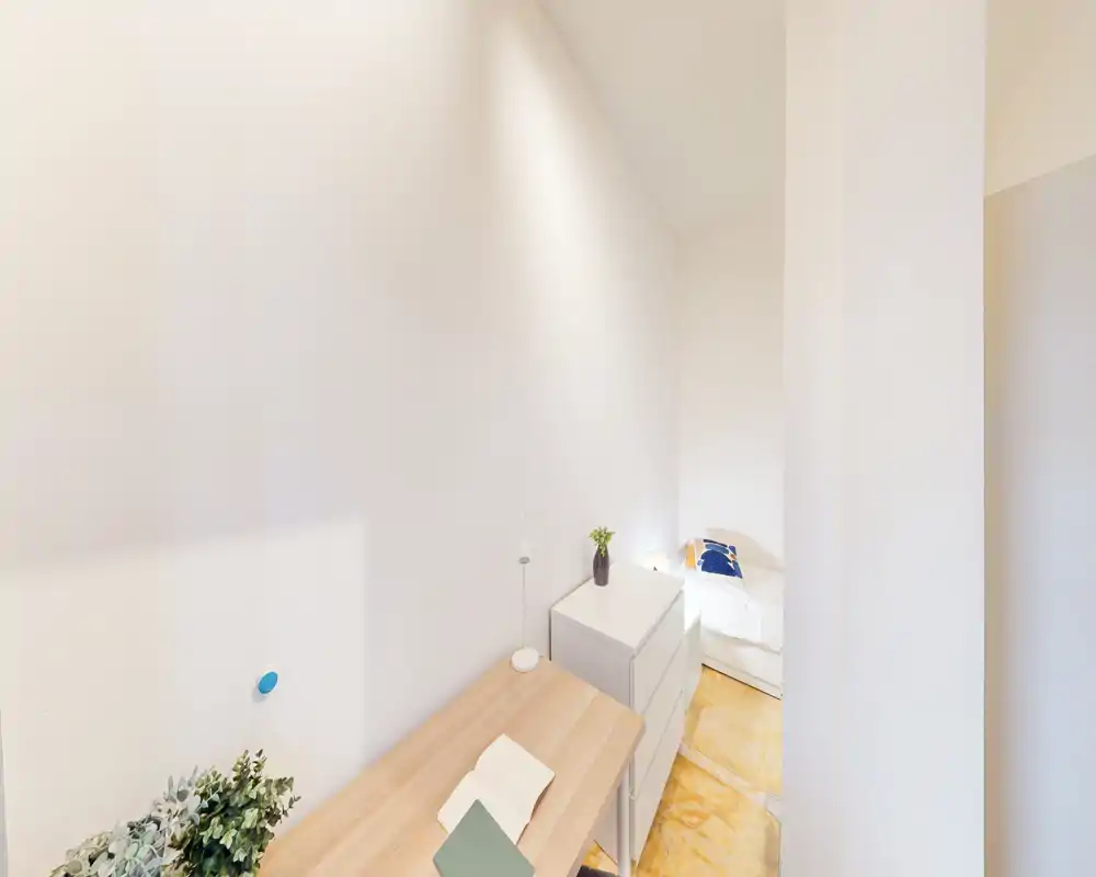 Small bright bedroom with a single bed, white walls, a chest of drawers and a wooden desk — minimal, modern and well lit.