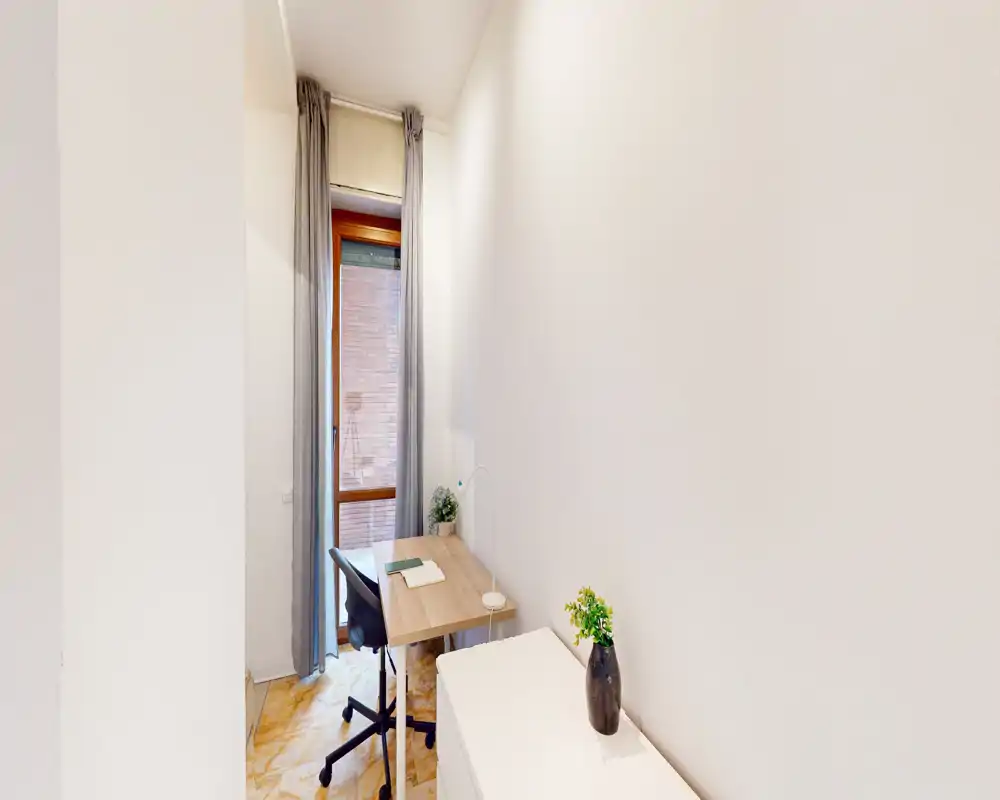 Compact home-office corner with a narrow desk, ergonomic chair, desk lamp and small plants next to a tall window providing natural light. Minimal, tidy and functional workspace ideal for remote work.