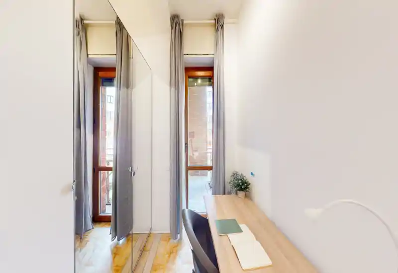 Small bright living area with a narrow desk and chair beside a tall window/door; neutral decor, curtains and a small plant create a simple, welcoming corner.