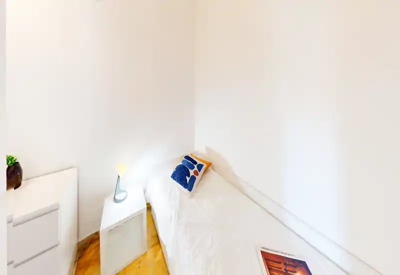 Small, bright single bedroom with a neatly made bed pushed into a corner, a decorative cushion, bedside lamp and clean white walls giving a minimalist, airy feel.