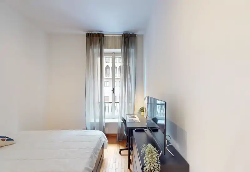 Bright, minimalist bedroom with a double bed, desk by the window and a TV on a low console. Natural light filters through sheer curtains, creating a clean and airy atmosphere ideal for short-term rentals.