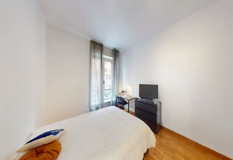 Bright, simply furnished bedroom with a double bed, bedside cushions and a desk by the window. Natural light and neutral walls create a calm, welcoming atmosphere.