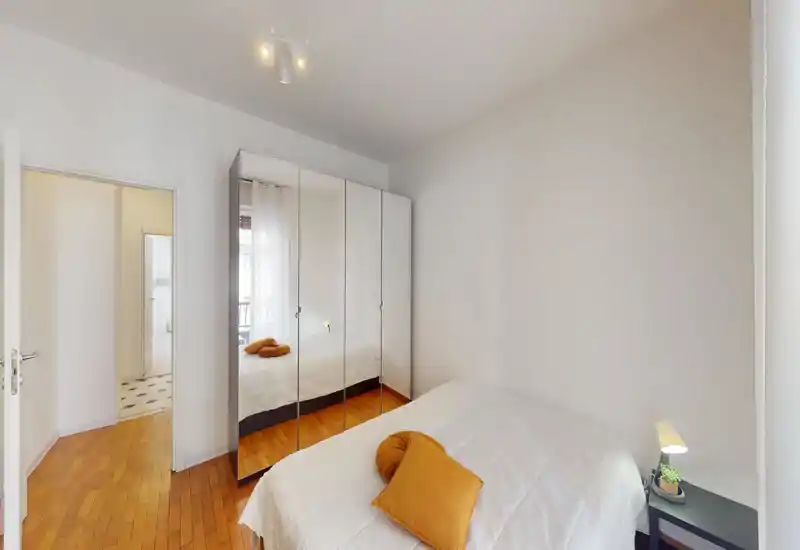 Dormitorio luminoso y minimalista con cama doble vestida en blanco, suelo de madera cálida y un armario con espejos que amplía el espacio. Lámpara de mesita y cojines ocre aportan calidez y un aire moderno — perfecto para anuncios de alquiler que destaquen un dormitorio limpio y luminoso.