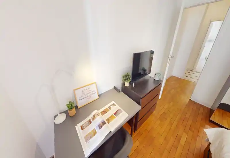 Rincón de dormitorio compacto con un pequeño escritorio, lámpara y cómoda con TV; vista parcial de la cama a la derecha. Espacio luminoso y ordenado con suelo de madera y decoración moderna.