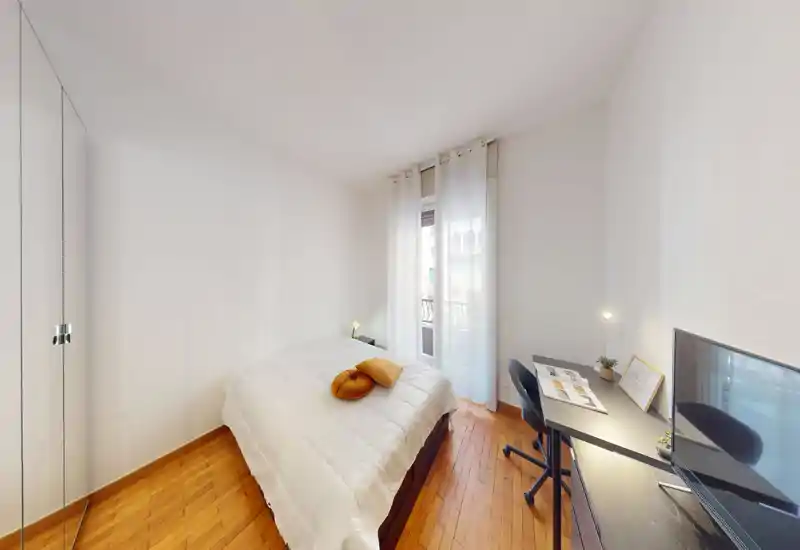Dormitorio luminoso y de estilo sencillo con cama doble, ropa de cama confortable y un pequeño escritorio junto a la ventana, perfecto para trabajar desde casa. La luz natural atraviesa las cortinas translúcidas sobre el suelo de madera.