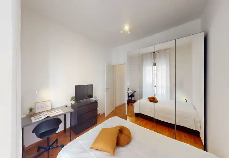 Dormitorio moderno y luminoso con cama doble, armario con espejo y un pequeño escritorio. Suelo de madera y detalles cálidos que crean un ambiente acogedor.