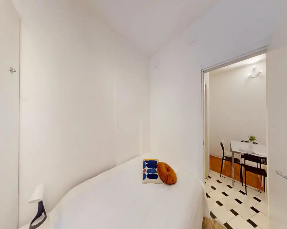 Camera da letto compatta e luminosa con letto matrimoniale ben rifatto, cuscini decorativi e pareti bianche minimal; la porta si apre su una zona pranzo.