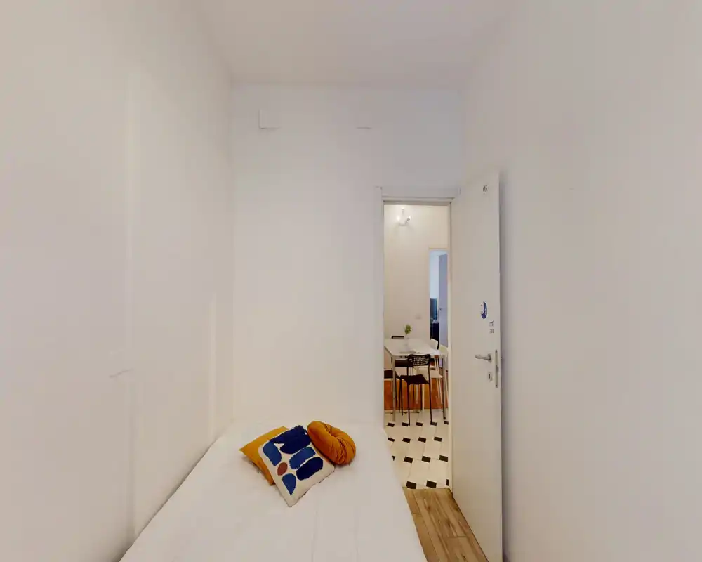 Camera da letto singola compatta e luminosa con letto ben rifatto e cuscini decorativi; pareti bianche che conferiscono un aspetto minimal e pulito, con l'ingresso verso la zona pranzo.