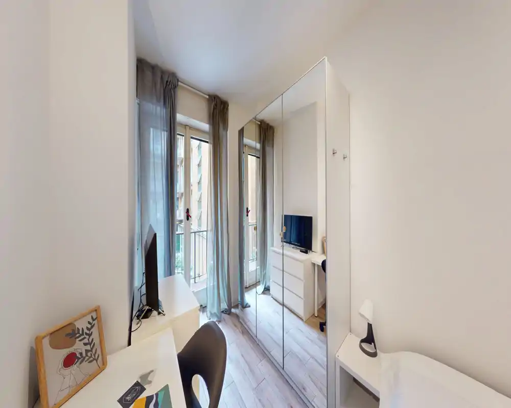 Angolo camera compatto e luminoso con una scrivania, un armadio con ante a specchio e un piccolo scorcio del letto. Tende leggere e la porta sul balcone offrono una luce naturale piacevole.