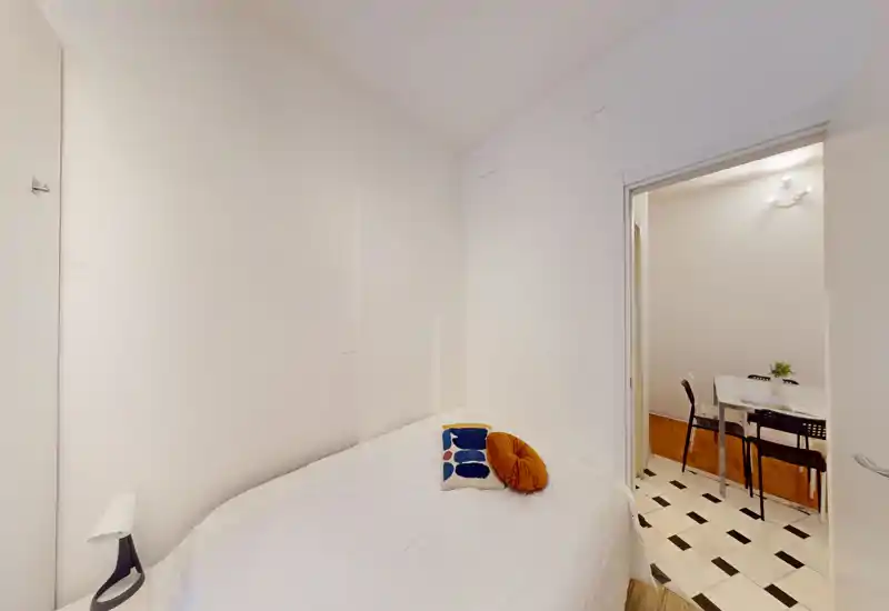 Camera da letto compatta e luminosa con letto matrimoniale ben rifatto, cuscini decorativi e pareti bianche minimal; la porta si apre su una zona pranzo.