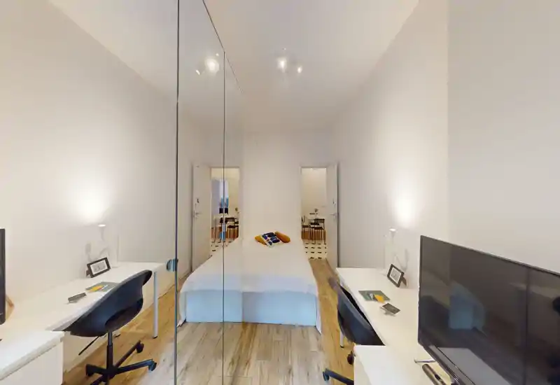 Camera da letto luminosa e minimal con letto al centro, armadio con ante a specchio che riflette la stanza e due scrivanie ai lati — pavimento in legno chiaro e cuscini decorativi per un’atmosfera rilassante.