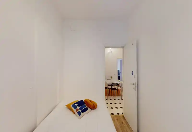 Petite chambre simple et lumineuse avec un lit bien dressé et des coussins décoratifs ; murs blancs pour une ambiance minimaliste, la porte donne sur la salle à manger.