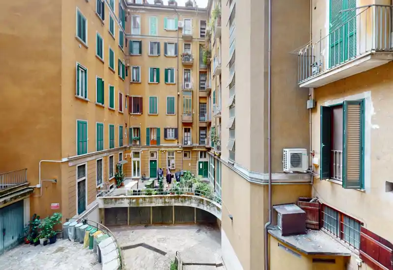 Vue sur une cour intérieure ensoleillée avec fenêtres d’appartements et petits balcons avec plantes ; idéal pour montrer le caractère de l’immeuble et l’espace extérieur commun.