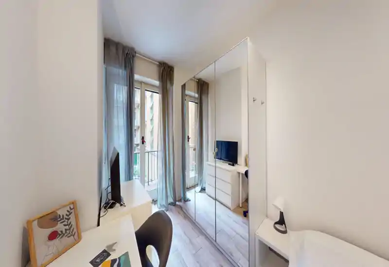Angolo camera compatto e luminoso con una scrivania, un armadio con ante a specchio e un piccolo scorcio del letto. Tende leggere e la porta sul balcone offrono una luce naturale piacevole.