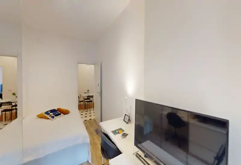 Camera compatta con letto singolo, cuscini decorativi e una piccola scrivania con TV — luminosa, pulita e arredata in modo semplice per soggiorni brevi.