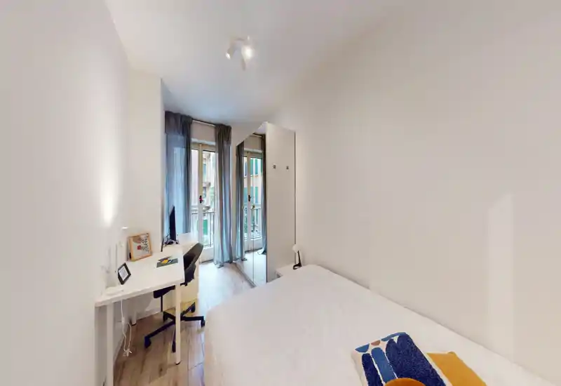 Camera da letto compatta e luminosa con letto matrimoniale, scrivania e accesso al balcone; arredi minimal e pavimento in legno chiaro creano un senso di ampiezza.