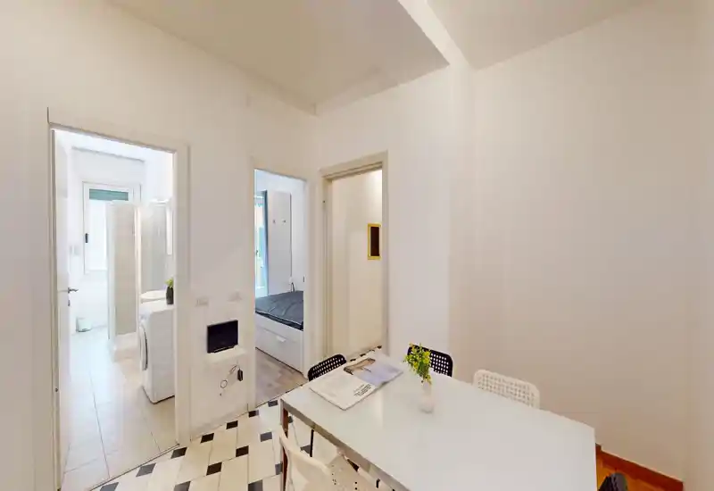 Espace séjour/salle à manger lumineux et moderne avec table blanche et chaises, décoration épurée et accès visible aux pièces adjacentes (salle de bain et chambre).