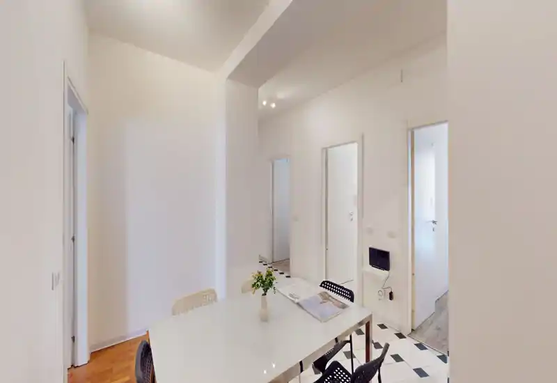 Espace repas minimaliste avec une table blanche, quatre chaises modernes et un petit vase ; pièce lumineuse et épurée donnant sur plusieurs portes.