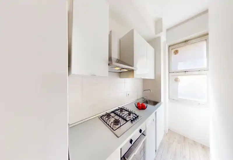 Cuisine moderne compacte et lumineuse avec plaque à gaz, hotte, évier et meubles blancs ; la lumière naturelle venant de la fenêtre met en valeur les surfaces nettes et le sol effet bois.
