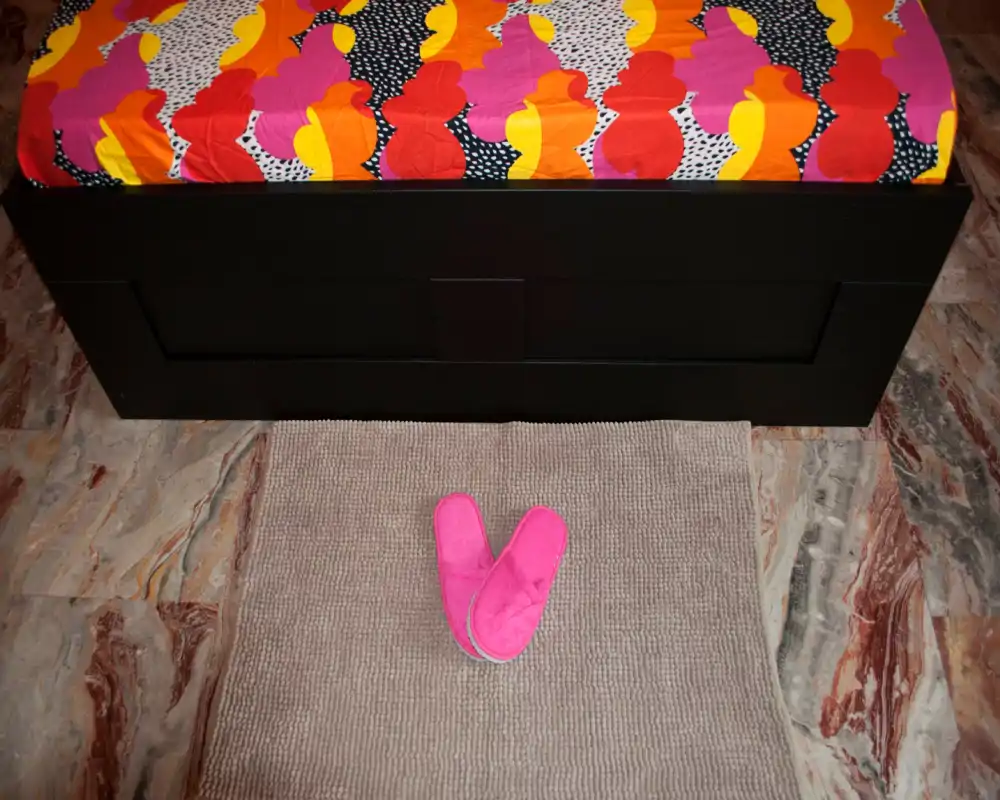 Angolo camera da letto luminoso e colorato con un letto singolo visibile coperto da una coperta vivace e un tappetino con pantofole rosa davanti: ambiente semplice e accogliente, ideale per un soggiorno confortevole.