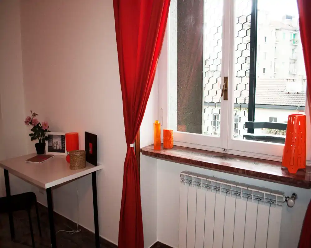 Angolo stanza luminoso e ben curato con scrivania e ampia finestra incorniciata da tende rosse, accessori caldi e termosifone sotto il davanzale.