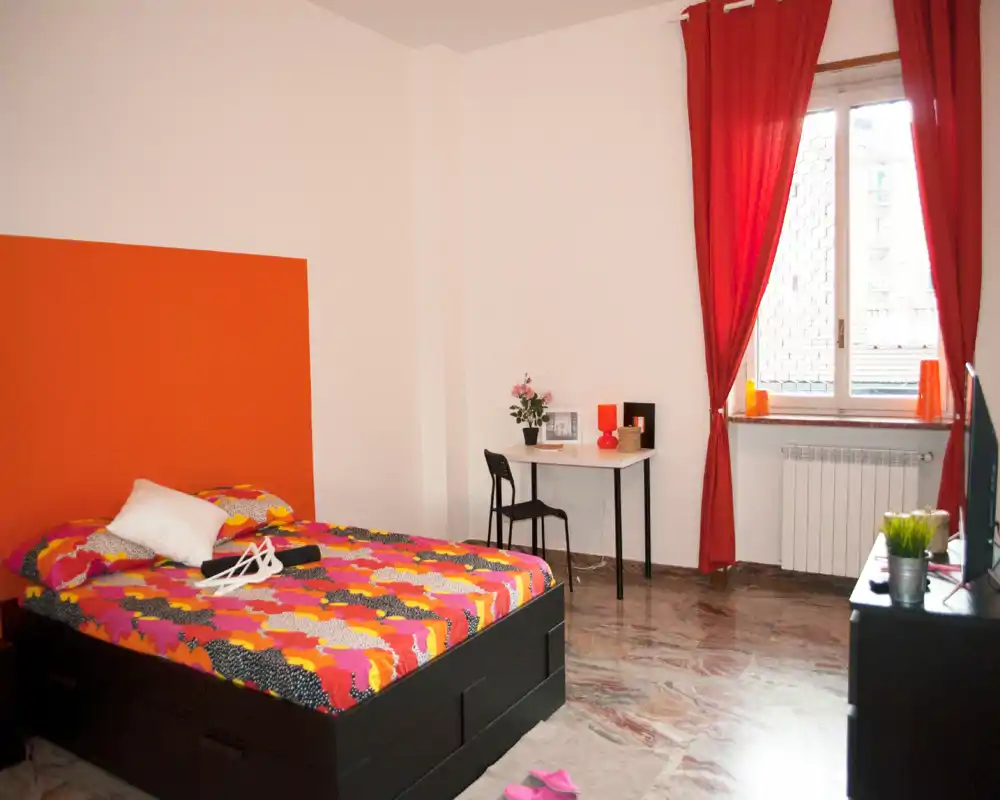 Camera da letto luminosa e colorata con letto matrimoniale, una scrivania semplice vicino alla finestra e tende rosse che creano un'atmosfera calda.