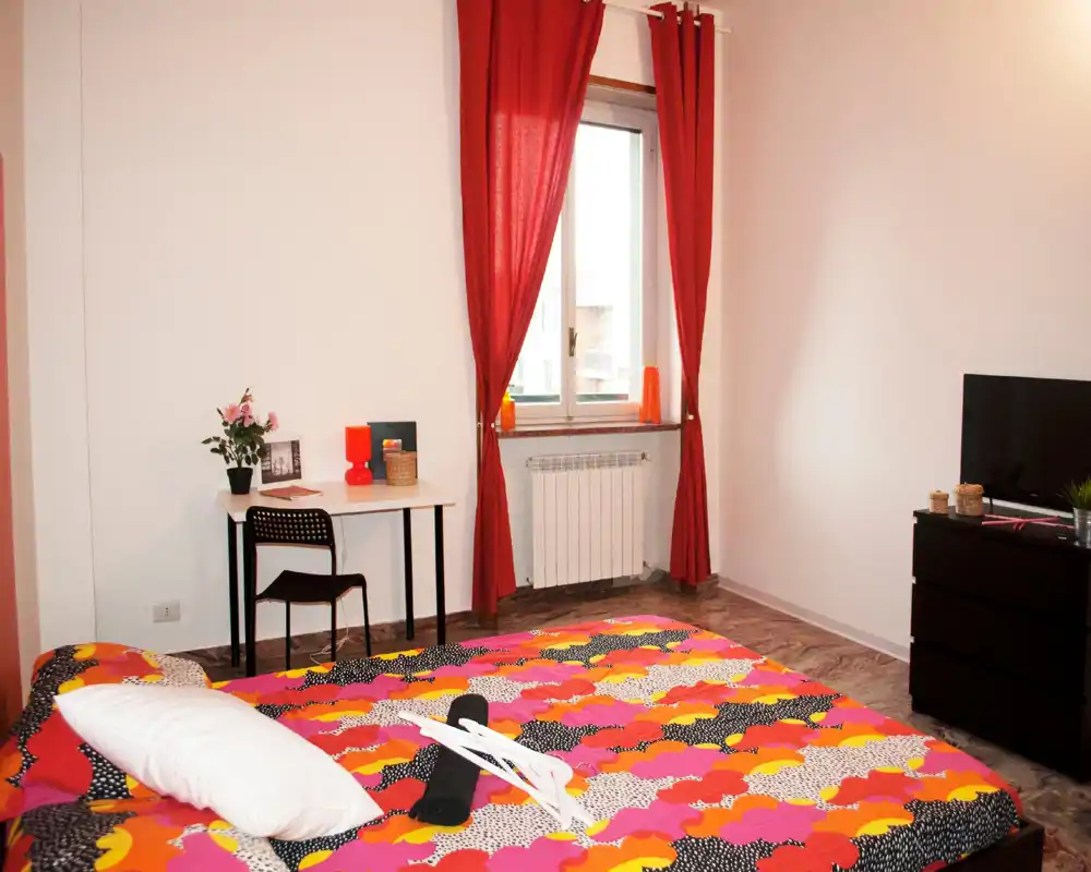 Camera da letto luminosa e colorata con un ampio letto in primo piano, decorazioni sul comodino, una scrivania vicino alla finestra e un comò con TV — ben allestita per annunci.