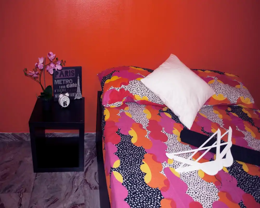Camera da letto luminosa e colorata con letto matrimoniale a fantasia, cuscino bianco e piccolo comodino con pianta decorativa e orologio — accogliente, ben illuminata e curata per le foto.