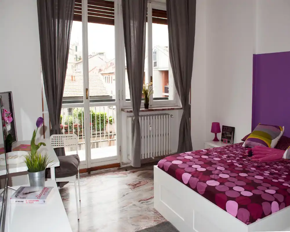 Chambre lumineuse et bien mise en scène avec un lit double aux draps violets à motifs, grande fenêtre avec rideaux laissant entrer la lumière naturelle, petite lampe de chevet et coin bureau — idéale pour un séjour urbain cosy.