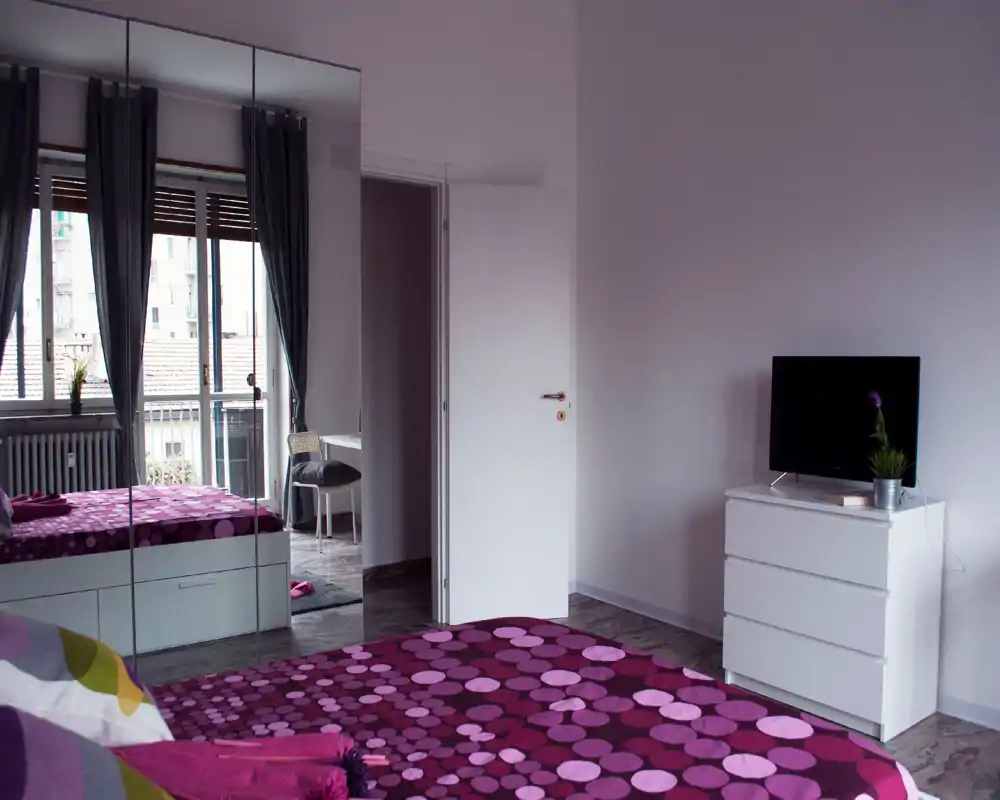 Chambre lumineuse et bien composée avec une couette violette à motifs, une armoire miroir et une commode avec télévision ; la lumière naturelle des grandes fenêtres apporte de l'air.