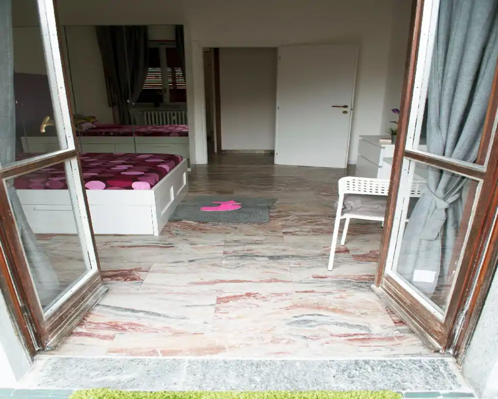 Chambre lumineuse vue par des portes ouvertes, avec un lit blanc et une couette rose à motifs, sol effet marbre et un petit coin de chevet — idéale comme chambre simple ou pour invités.