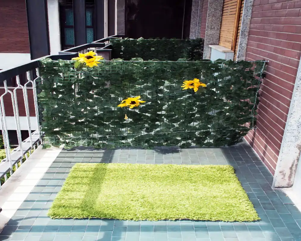 Petit balcon carrelé avec brise-vue végétal et tournesols artificiels, ainsi qu'un tapis vert ; espace extérieur lumineux et soigné.