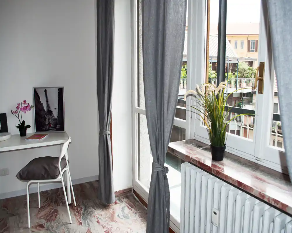 Coin de chambre lumineux avec grande fenêtre et jolis rideaux ; un petit bureau et des plantes décoratives offrent un espace de travail apaisant.