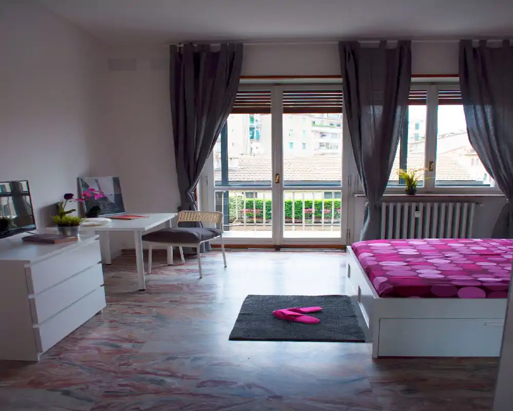 Chambre moderne et lumineuse avec un grand lit à motifs près d’une large fenêtre donnant sur le balcon. La pièce comprend un petit bureau, une commode et des textiles confortables, idéale pour un séjour agréable.